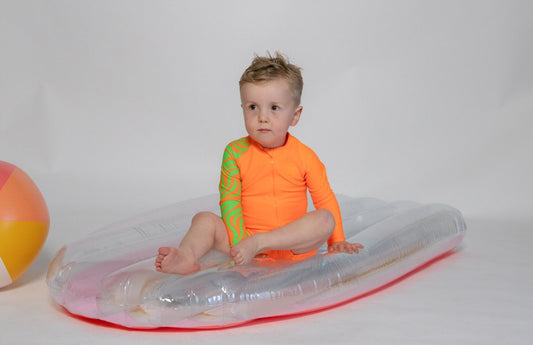 A toddler in a bright orange long sleeve swimsuit with a unique zebra print on the sleeves is sitting on an inflatable ring, with a ball in the background.